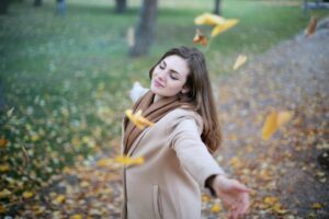 A young woman with long brown hair wearing a beige sweater and a brown scarf holding her arms out at shoulder level and eyes closed while standing on a leaf strewn path with grass in the background and yellow leaves falling