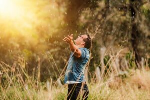 A man standing in a field near a forest in the rain with the sun in the background wearing a blue shirt and black pants with his arms outstretched and his head raised toward the sky