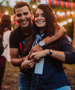 A young smiling latino man with his left arm around a young smiling latino woman who is holding both of his hands at an outdoor party