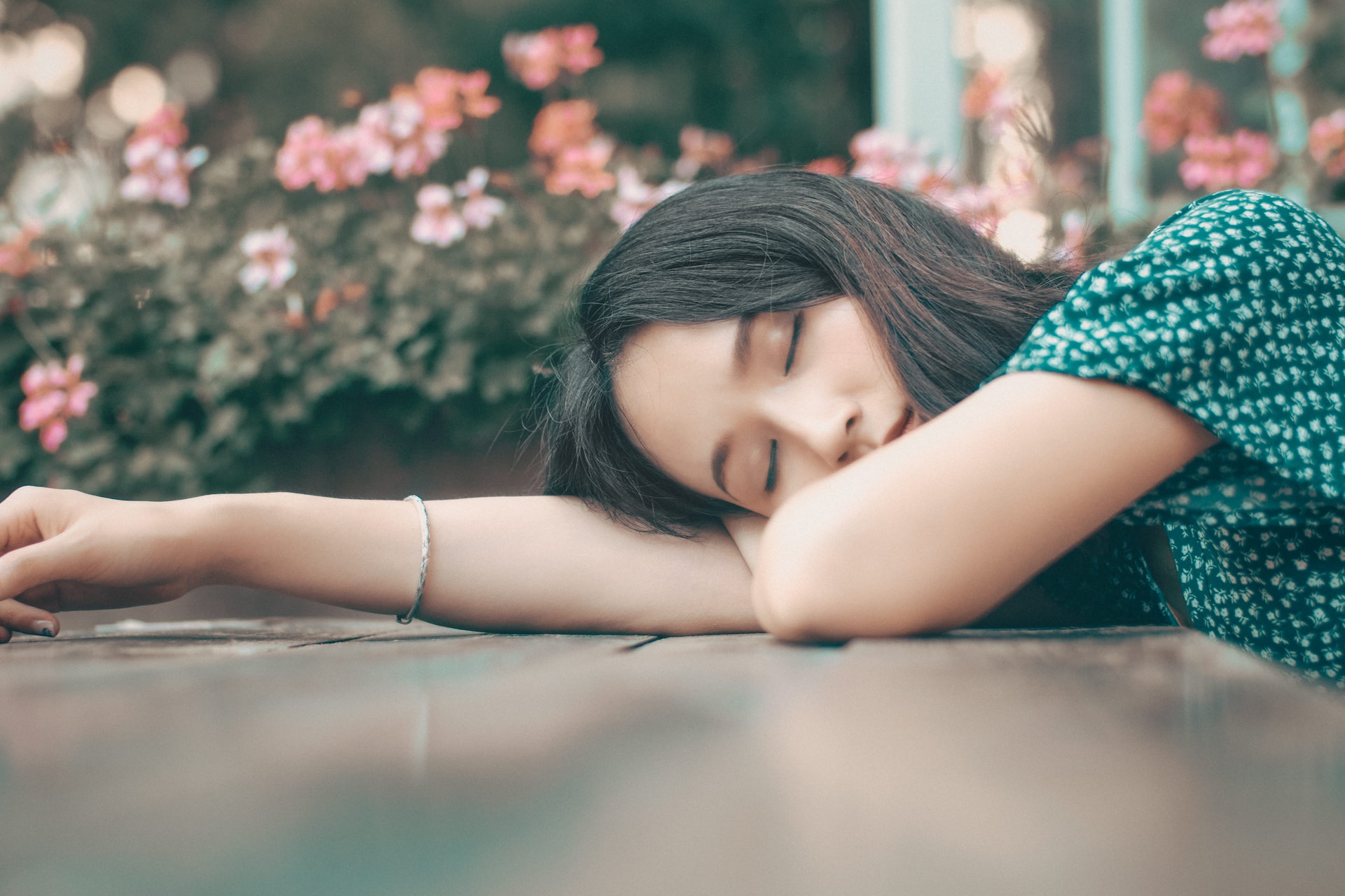 A young Asian woman asleep with her head resting sideways on one of her outstretched arms on a table with red and pink flowers in the background