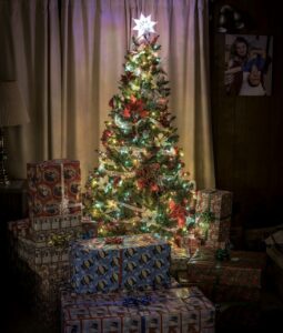 A decorated and lit Christmas tree with an assortment of wrapped gifts surrounding the base in the middle of the backside of a room in a home