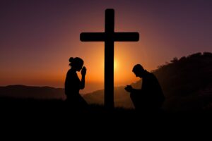 A silhouette of a man and a woman kneeling in prayer at a cross with a rising sun, hills, and trees in the background