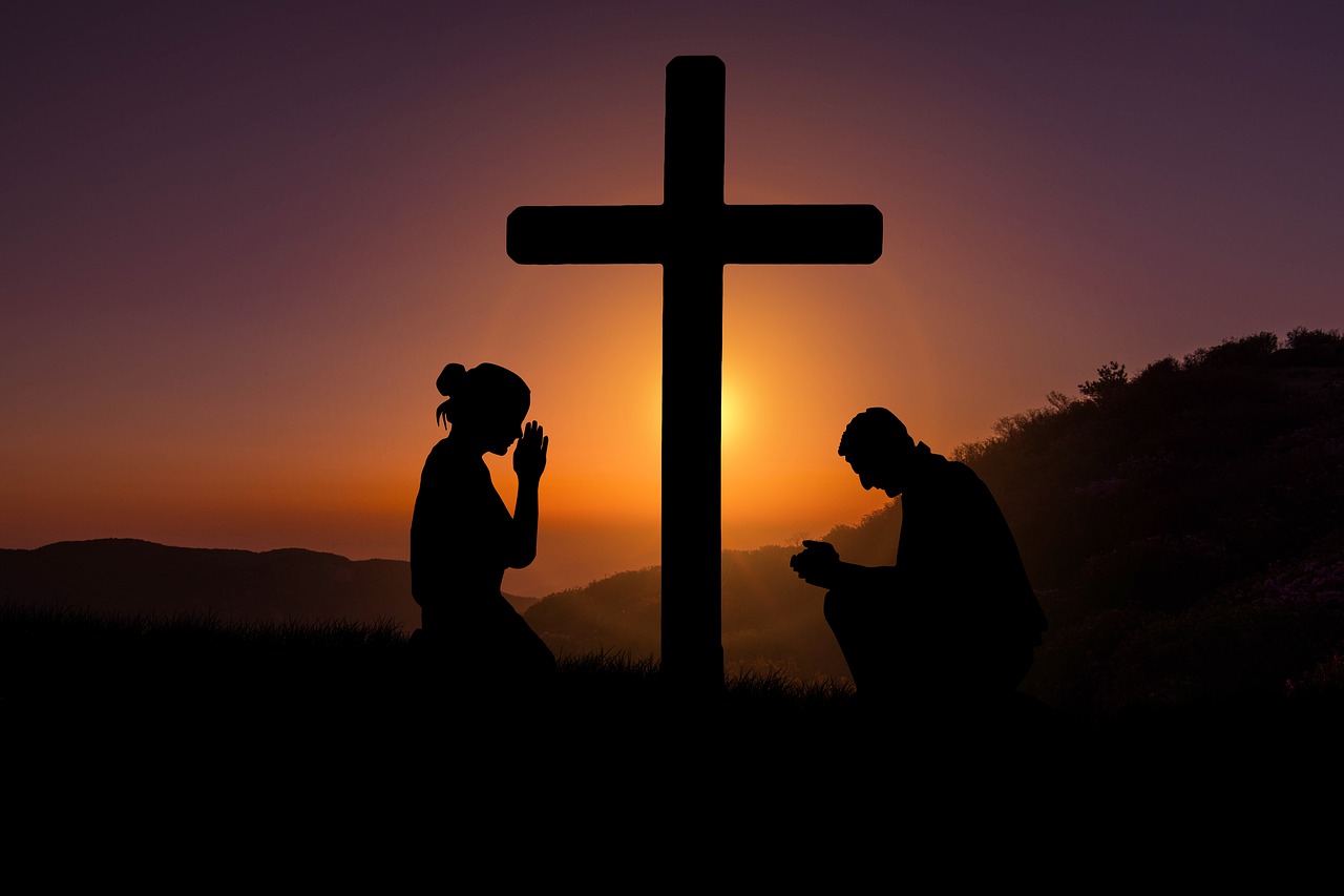 A silhouette of a man and a woman kneeling in prayer at a cross with a rising sun, hills, and trees in the background