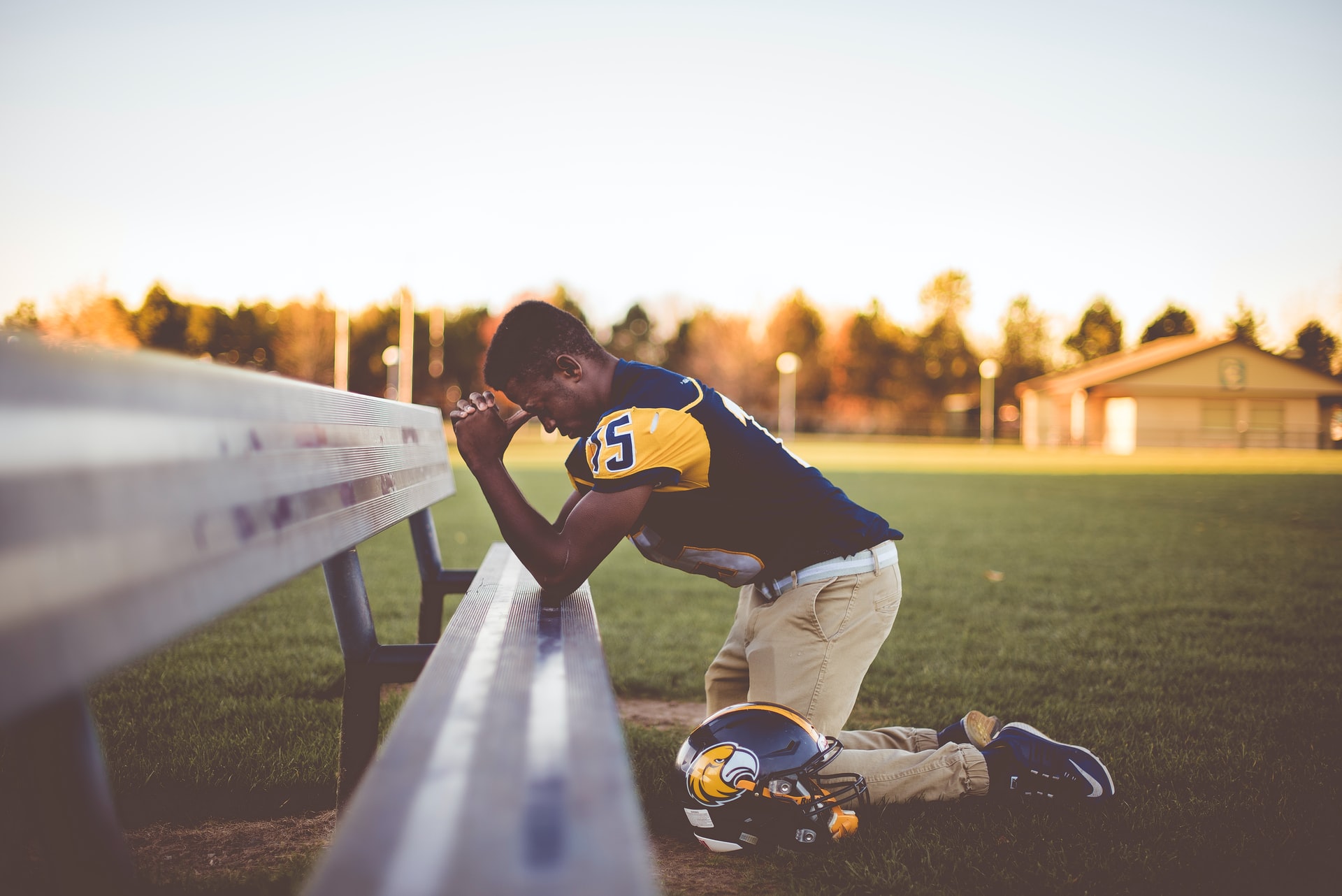 An African-American football player in full yellow and black uniform with tan pants and helmet near his leg kneeling in prayer on a metal bench with blue sky, sunlit trees and a building in the distant background