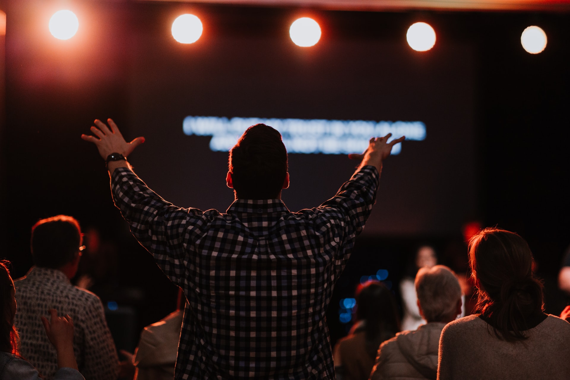 A photo of the backside of a man wearing a longsleeved plaid shirt standing with his arms in the air in praise among a large group of people seated in a darkened room with stage lights overhead and blurred words on a screen in the background