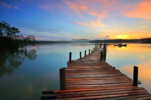 A long wooden dock on a still blue lake with a boat in the distance, mountains in the far background, various shades of orange in the sky and on the lake from a rising sun, with a cluster of trees on the far left