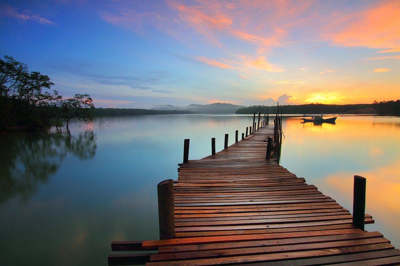 A long wooden dock on a still blue lake with a boat in the distance, mountains in the far background, various shades of orange in the sky and on the lake from a rising sun, with a cluster of trees on the far left