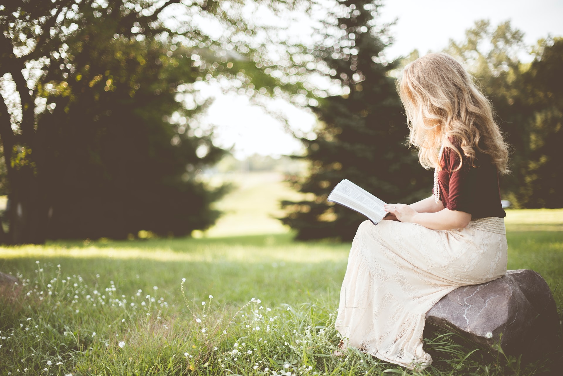 A blond haired Caucasian woman with a maroon shirt and a long white dress seated on a rock in a grassy area and trees in the background on a sunny day, reading a book