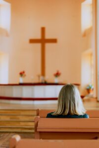 A photo of the back of a blond haired woman seated in a pew in a well lit church with a large brown cross in the background