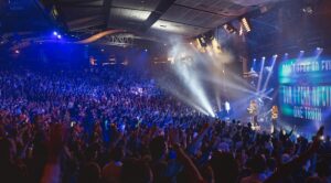 A photo of a large auditorium of a church with thousands of people standing with hands in the air in a darkened area with numerous stage lights highlight singers and instrumentalists on a stage and a inspirational slogan displayed on a large screen behind them