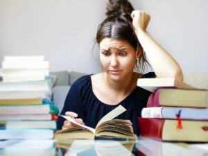 A young woman with black hair and a black short sleeved shirt sits at a table filled with stacks of books staring incredulously, with her left arm resting on a small pile of book and leaning on her head, at an open book laying flat in the table