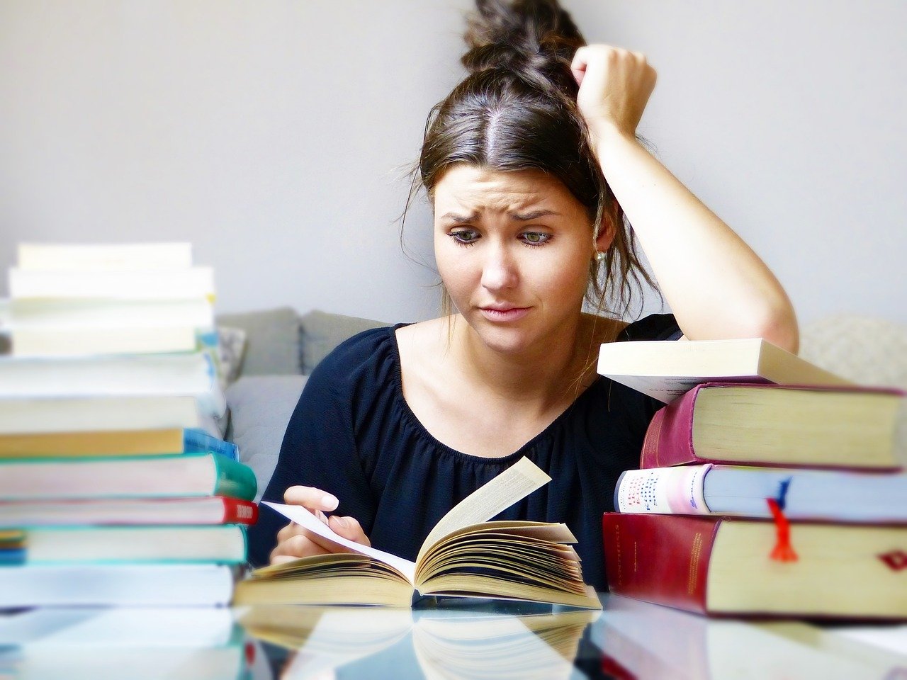 A young woman with black hair and a black short sleeved shirt sits at a table filled with stacks of books staring incredulously, with her left arm resting on a small pile of book and leaning on her head, at an open book laying flat in the table