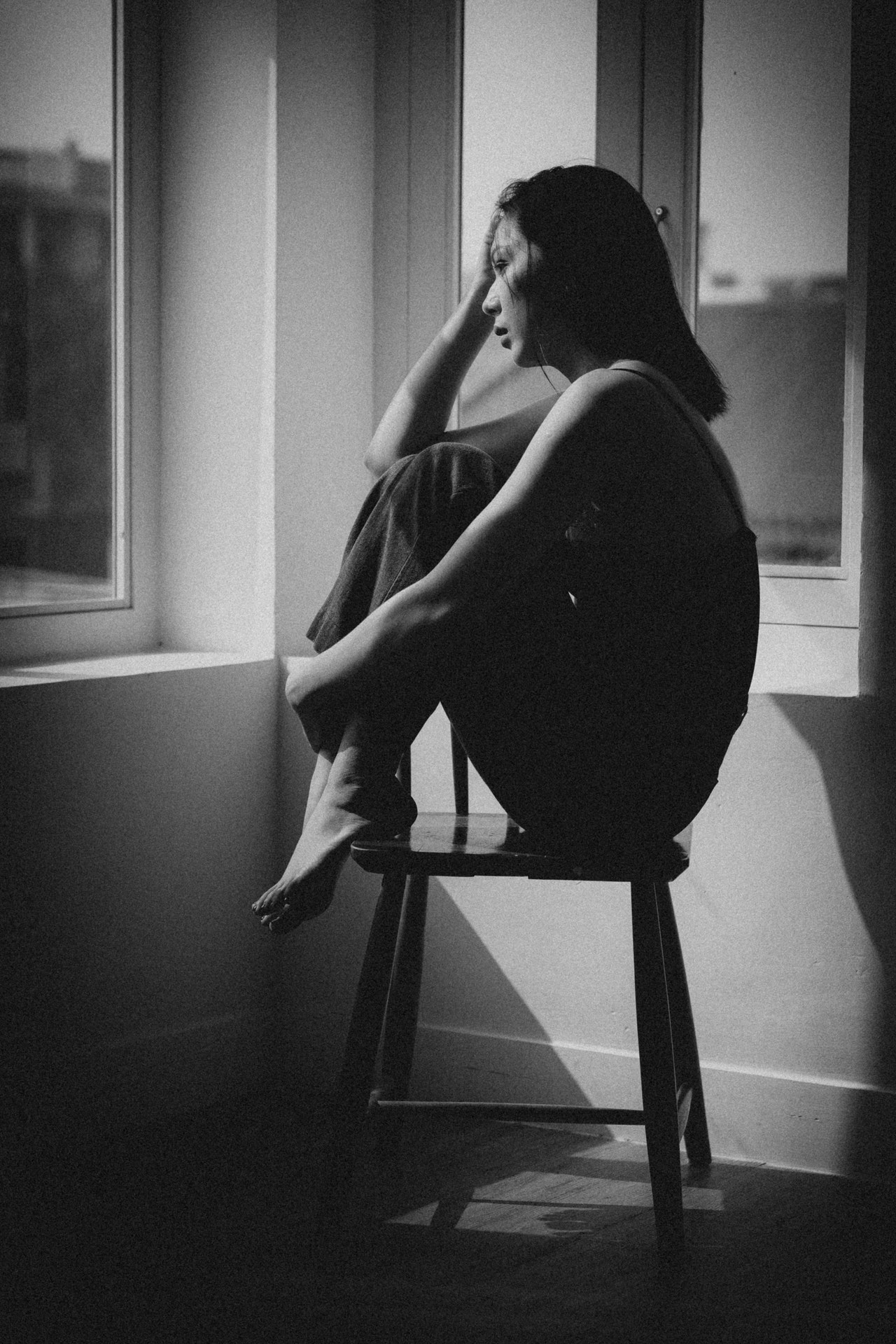A black & white photo of an Asian woman with a sad, distressed expression seated in the corner of a room between two windows in sunlight with her legs folded and feet resting on the chair with one arm holding her legs and the other raised holding her head