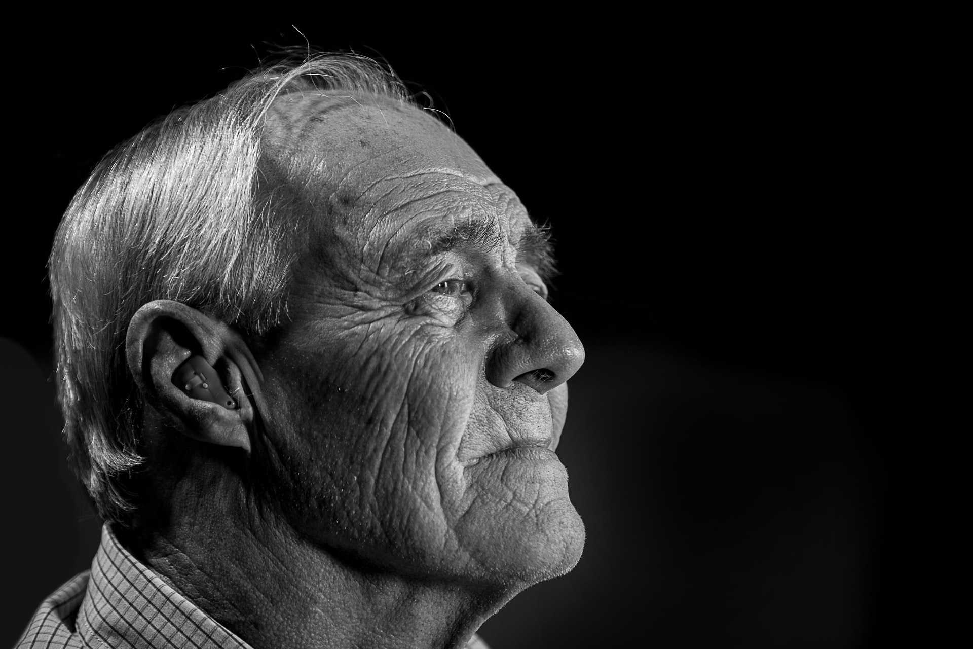 A black and white side portrait of an elderly gray haired caucasian man wearing a hearing aid on a black background reflecting on the love and happiness on his past.