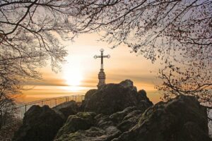 A metal cross on the rocky summit of Litermont in Germany with tree branches surrounding it and a hazy orange sunset in the background
