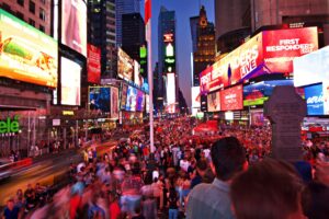A photo looking down Times Square in New York City at night with hundreds of people and brightly lit full colored electronic billboards on the buildings of both sides of the street