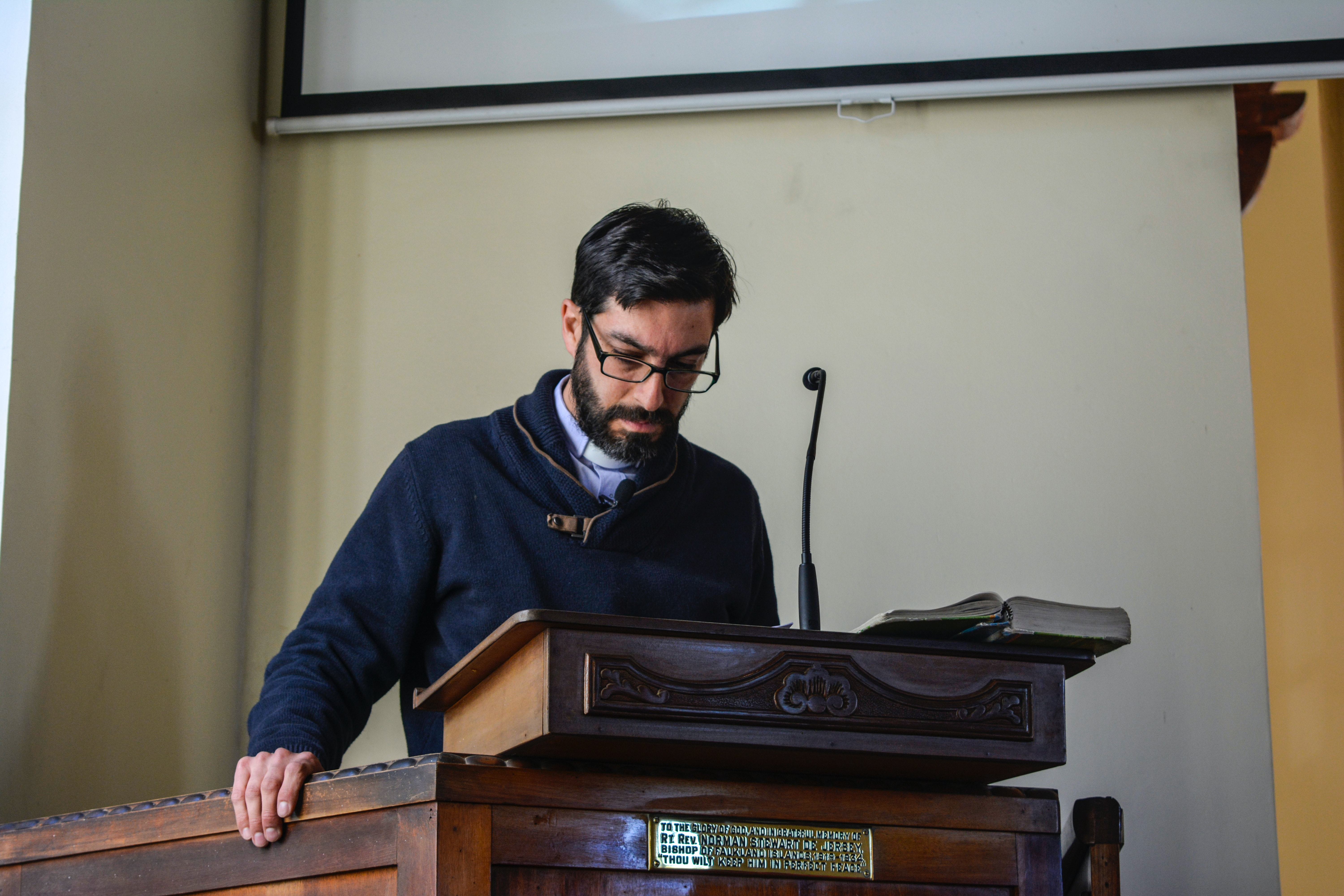A middle-aged, dark haired Caucasian man with a beard, mustache, and glasses looking down while standing behind a pulpit in a church