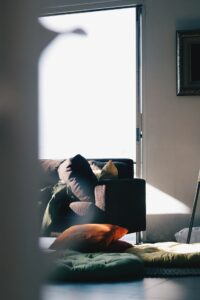 A photo of a sunlit room with a partial view of a dark couch and a person, mostly out of view, with a green shirt or jacket lying on it, colored pillows strewn on the floor, and a sliding glass door displaying an overexposed white sky in the background