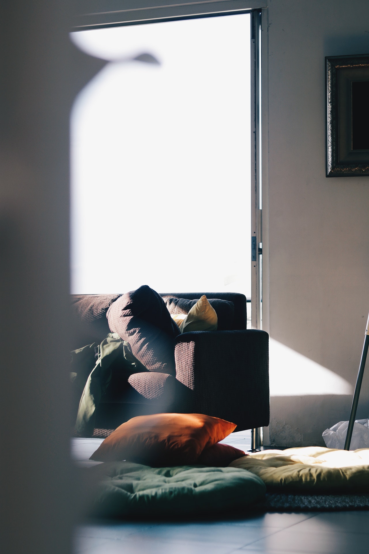 A photo of a sunlit room with a partial view of a dark couch and a person, mostly out of view, with a green shirt or jacket lying on it, colored pillows strewn on the floor, and a sliding glass door displaying an overexposed white sky in the background