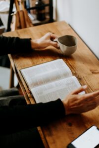 An open Bible laying flat on the surface of a small wood grain desk with a hand on left side holding a coffee cup and the other hand slightly raised in the air on the right side