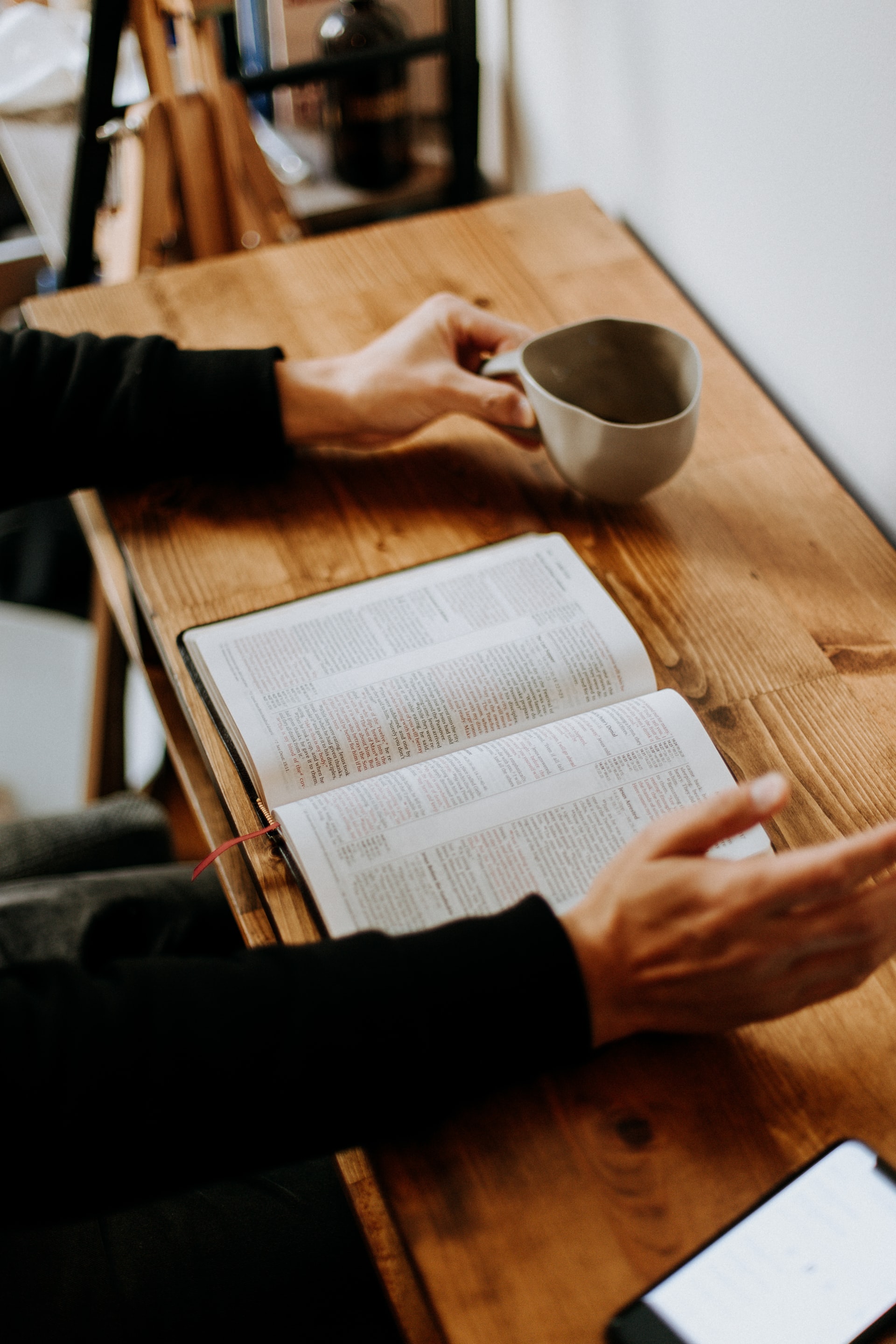 An open Bible laying flat on the surface of a small wood grain desk with a hand on left side holding a coffee cup and the other hand slightly raised in the air on the right side