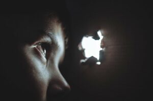 Boy looking though a small hole in a metal wall, with light shining on his face