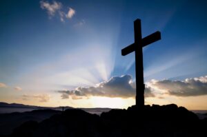 A silhouette of a large cross, on a rocky surface, on the right side of a photo with blue sky, assorted clouds, a setting sun with beams protruding out