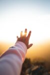 A Caucasian man's hand, with arm in a pink long sleeved shirt, raised towards a bright setting sun with part of the sun coming though his fingers