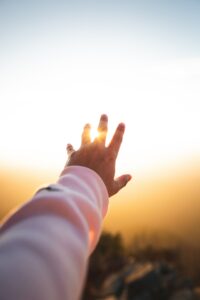 A Caucasian man's hand, with arm in a pink long sleeved shirt, raised towards a bright setting sun with part of the sun coming though his fingers