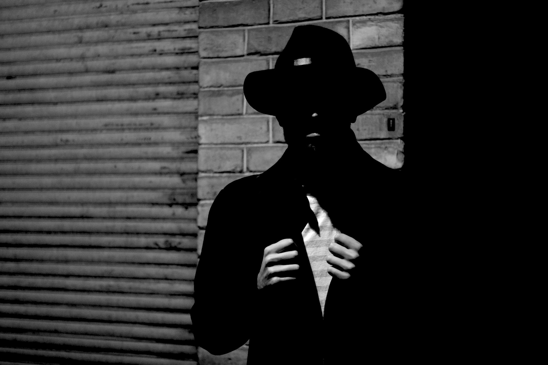 A black and white photo of a man wearing a black narrow brimmed hat, dark jacket, partially exposed white striped shirt with hands pulling it open while standing against a brick wall next to a corrugated roll-up door