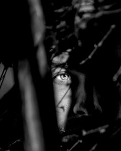 Black and white photo of a Caucasian man's face hidden by various shadows with only his eye and cheek partially exposed in the light