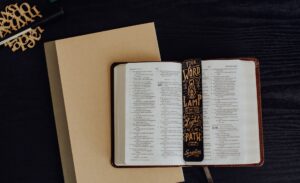 A open Bible and a black with gold scripture bookmark laying flat on top of a tan notebook on a black surface