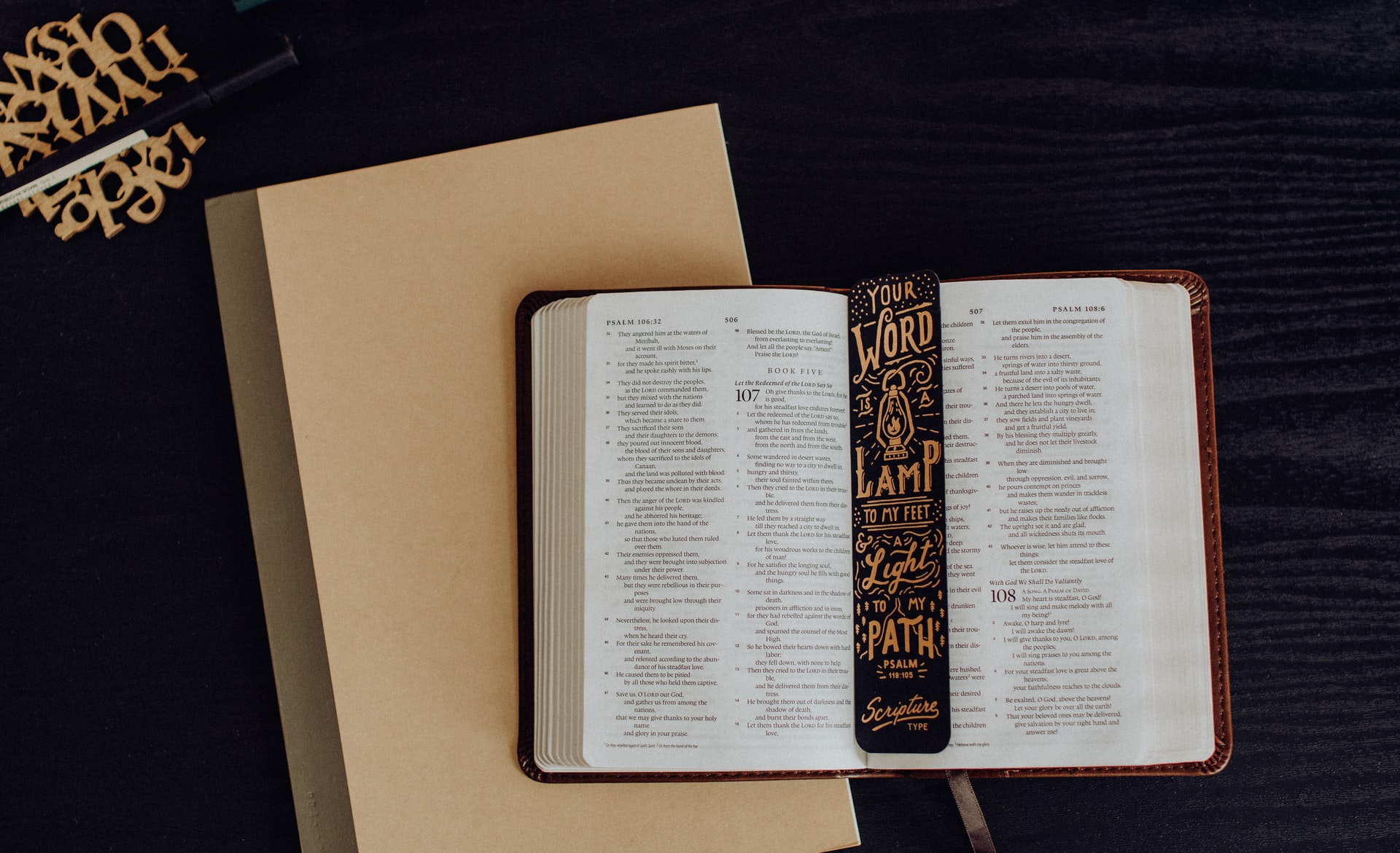 A open Bible and a black and gold bookmark laying flat on a tan notebook on a black surface symbolizing when seeking your blessings the need for the emphasis of God's Word 