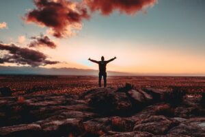 A silhouette of the backside of a man with arms raised while standing on several large rocks amidst a large desert-like area covered in a reddish hue with a setting sun and assorted clouds in a portion of the sky overhead in the same color as the ground
