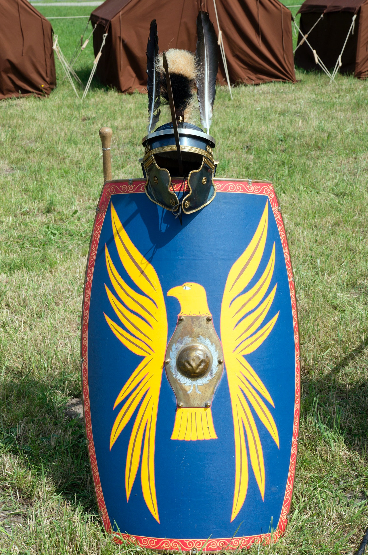 Photo of a blue Roman/medieval shield with a red decorative border and a large yellow eagle crest resting on the ground with a helmet attached to the top and brown tents in the background