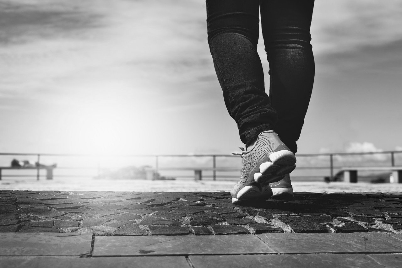 A black and white photograph of a person in dark pants wearing athletic shoes walking on a stone inlaid platform with bright sun and and a railing in the distance