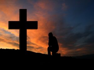 A silhouette of a man kneeling at a large cross against an orange colored clouded sky at sunset