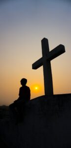 A silhouette of a person seated at the base of a large cross on a hill with a hazy yellow sun setting in the background