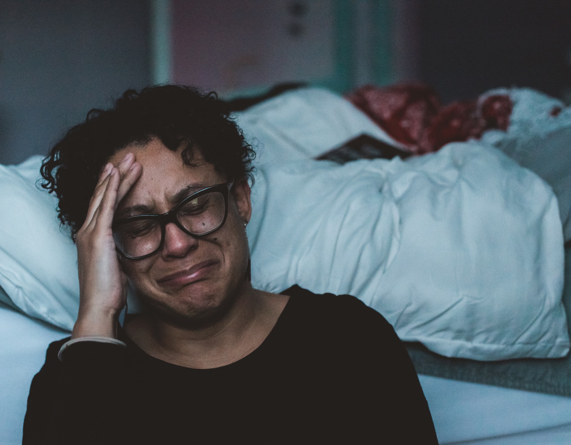 An older Hispanic woman with glasses sitting on the floor against the side of her bed in tears while holding her right hand against the side of her face while struggling with difficult situations