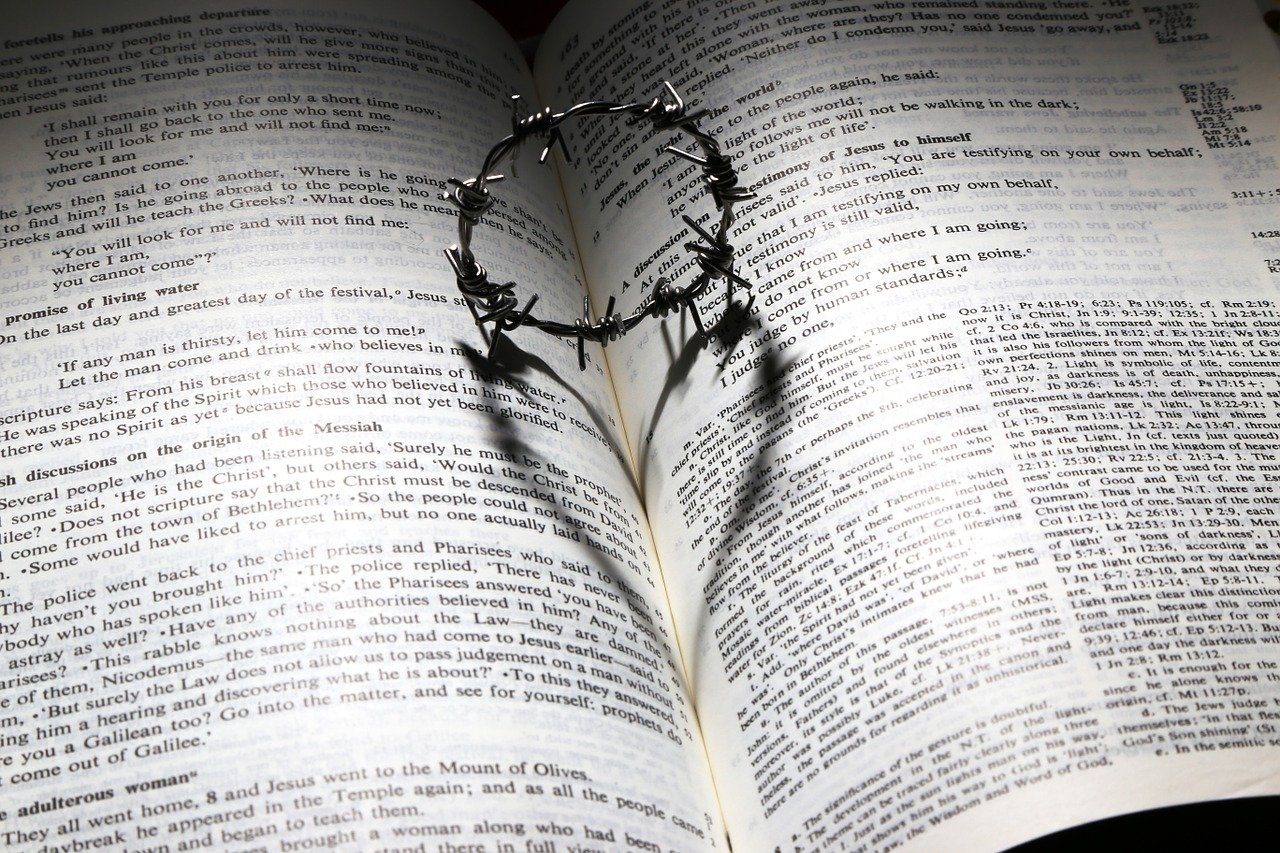A small ring of barbed wire resting vertically in the fold of an open Bible casting a heart shaped shadow