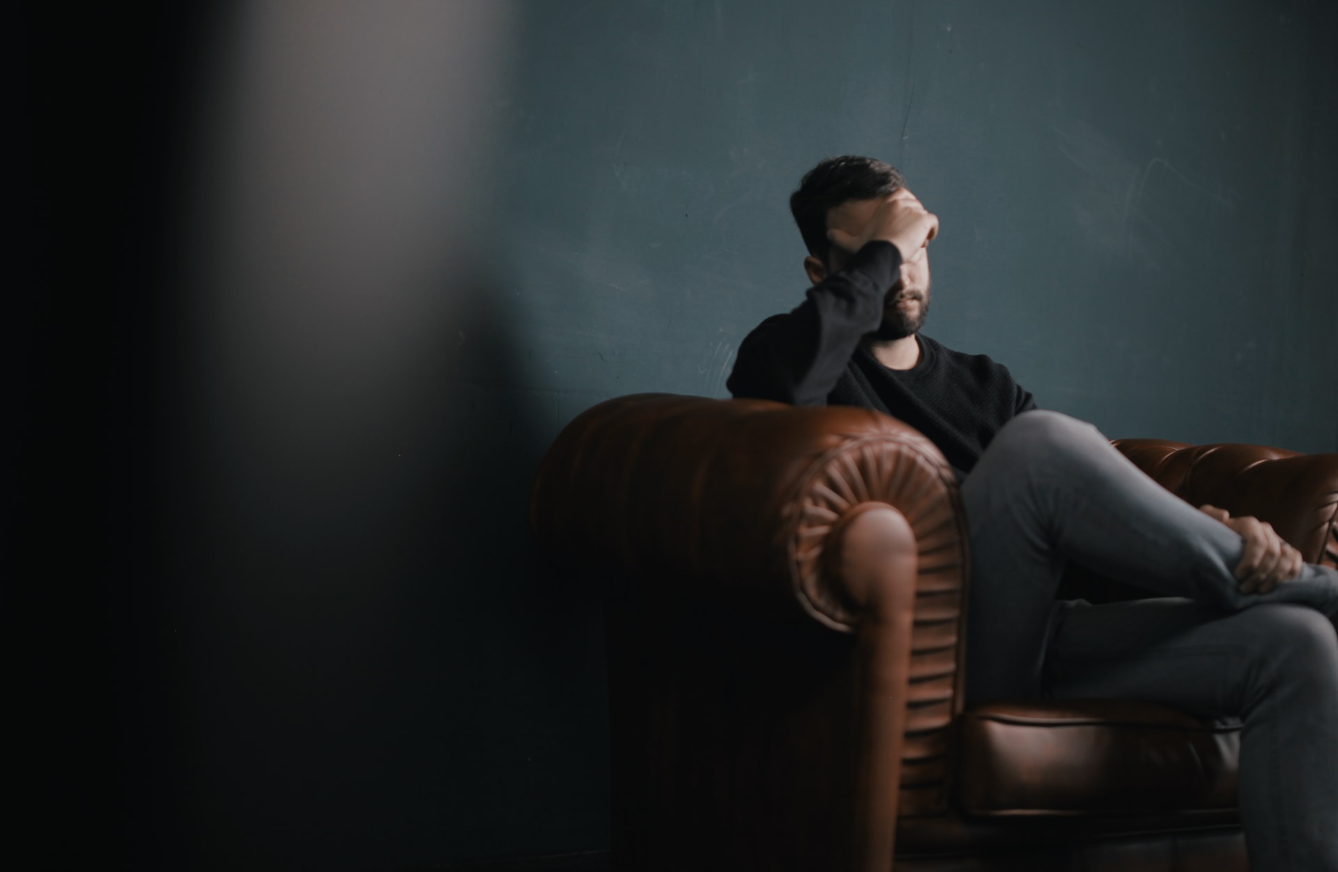 A middle aged Caucasian man sitting on the left side of a brown leather couch in the corner of a low lit room with a dark green wall, holding his hand over his forehead with his leg crossed