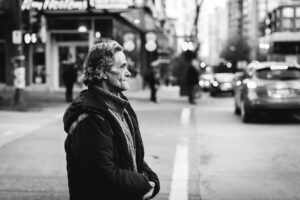 A black and white photo of an older Caucasian man wearing a sweater and dark coat standing near the edge of a street in the downtown region of a city waiting to cross to the other side
