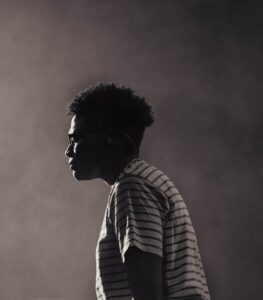 A black and white photographic portrait of a man of African descent wearing a striped white shirt and glasses with his eyes closed standing against a plain background