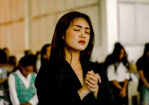 A young Asian woman standing with her eyes closed in prayer with others in the background doing the same