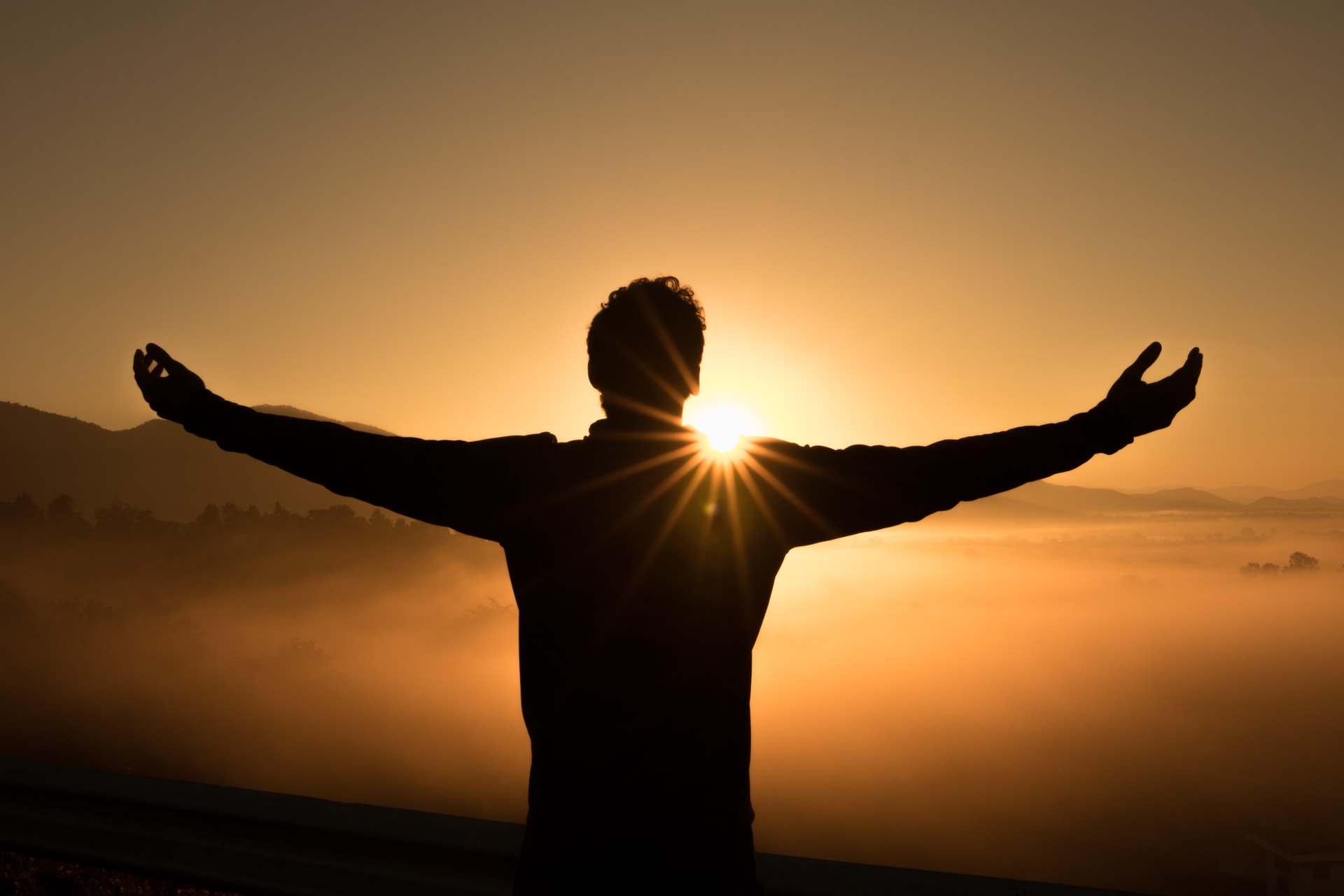 A silhouette of a the backside of a man standing with his arms outstretched and a setting sun just over his shoulder and a orange tinted foggy valley below