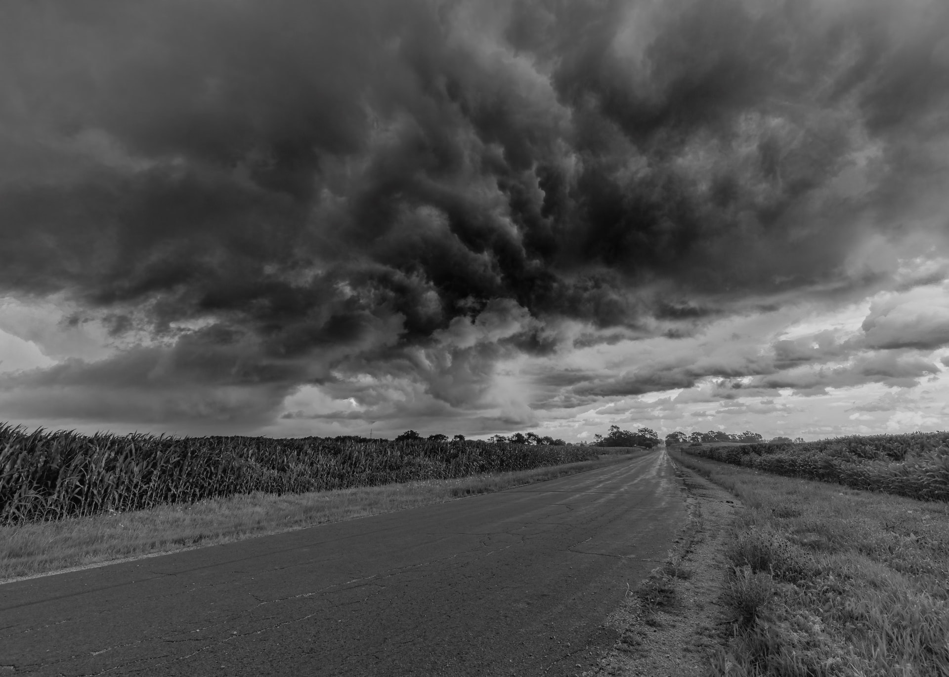 A black and white photo of approaching storm clouds alongside a small two-lane rural road between two sections of farmland