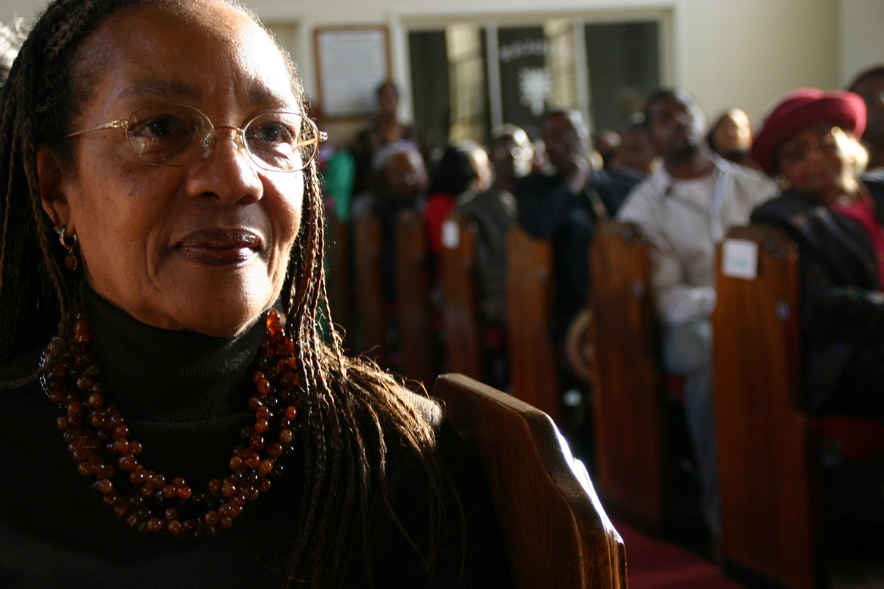 A photo of an older Afro-American woman seated in a pew during a church service with other congregants in the background to her right