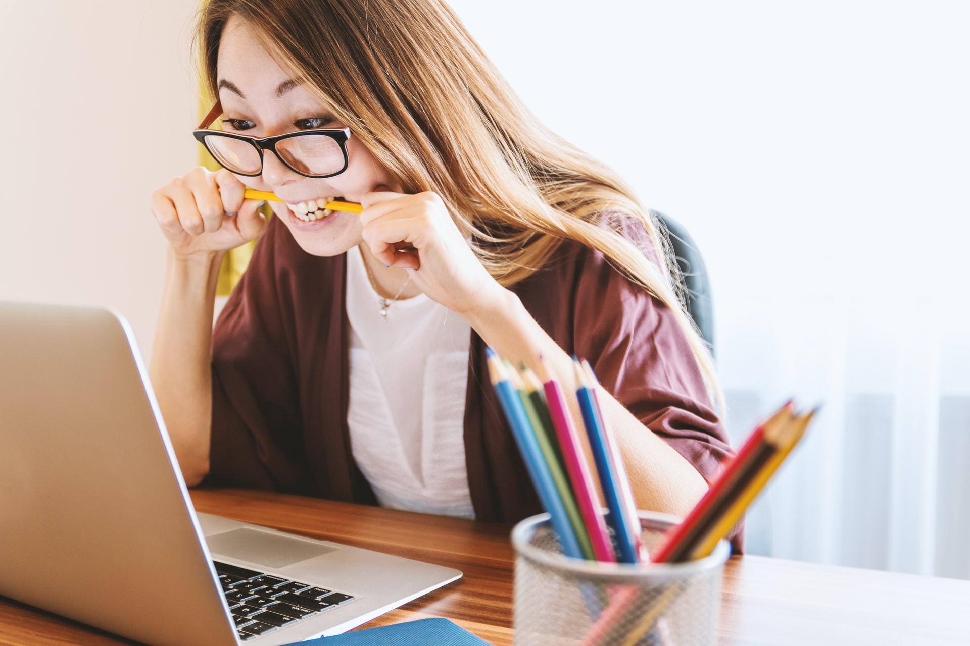 A young Caucasian/Asian woman with long light brown hair wearing large glasses seated in front of a bright white background and leaning slightly over a laptop computer while holding a pencil with both hands between her clenched teeth showing a lack of patience