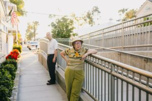 An elderly woman wearing a hat and a light green striped shirt with a single yellow flower and olive green slacks facing slightly toward the camera while leaning on a railing of a disability ramp in a sunlit suburban community with an elderly man standing behind her in a white shirt and dark pants and a somewhat incredulous expression on his face