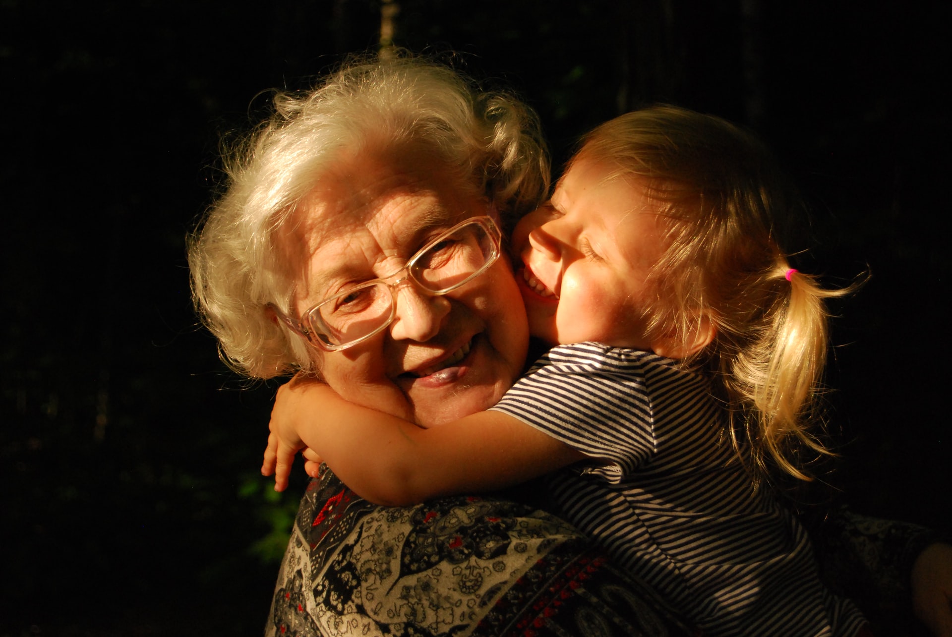 A head shot portrait showing the love and devotion of a smiling elderly woman with glasses and white hair holding a blond-haired young girl in her arms with the girl's arms wrapped around her neck with sunlight streaming on both of them